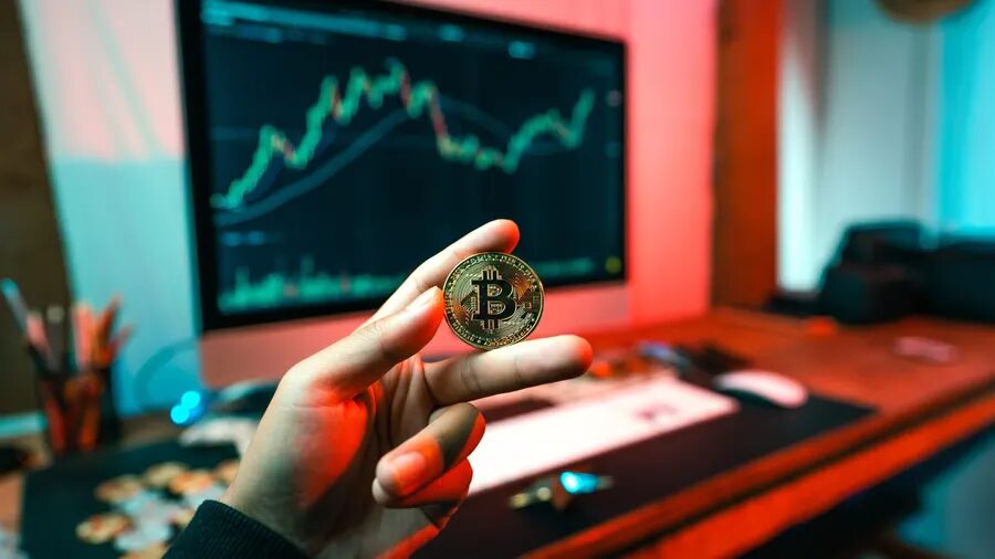 man holding bitcoin in front of a TV monitoring a trade chart