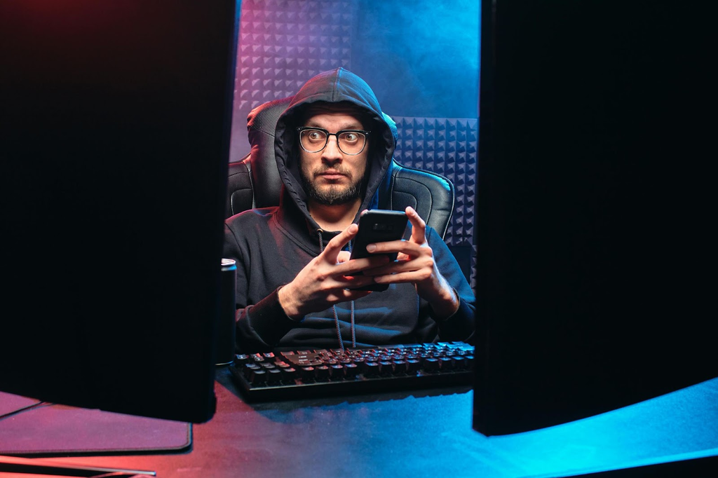 Een man met een angstaanjagende blik die op een computer werkt en op zijn mobiele telefoon drukt