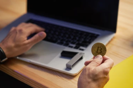 Une main tenant un bitcoin, l'autre main posée sur l'écran d'un ordinateur portable. 