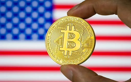Close-up of a hand holding a gold Bitcoin coin in front of a blurred United States flag.