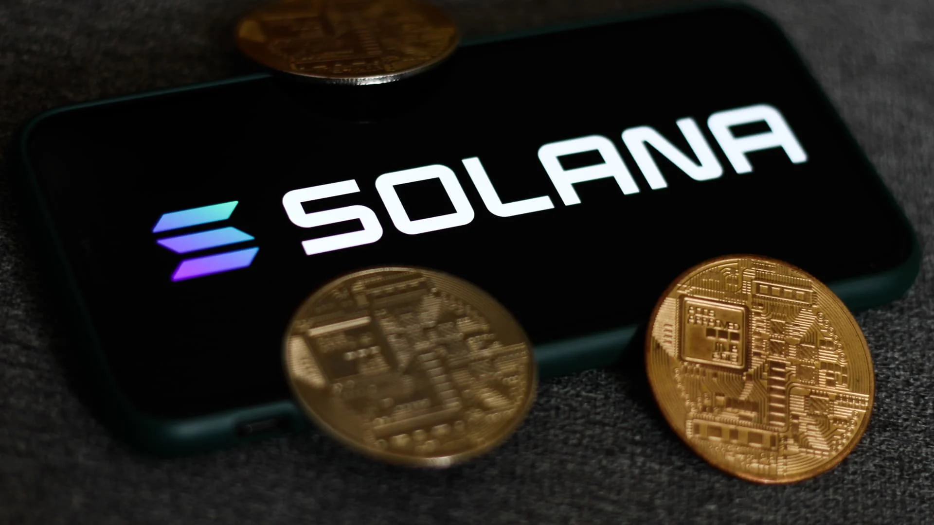 Smartphone displaying the Solana logo and name on its screen, surrounded by gold cryptocurrency coins on a dark surface.