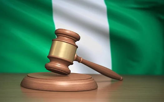 Ein hölzerner Richterhammer ruht auf einem Klangblock vor der nigerianischen Flagge und symbolisiert Recht, Verordnung und Rechtsweg in Nigeria.