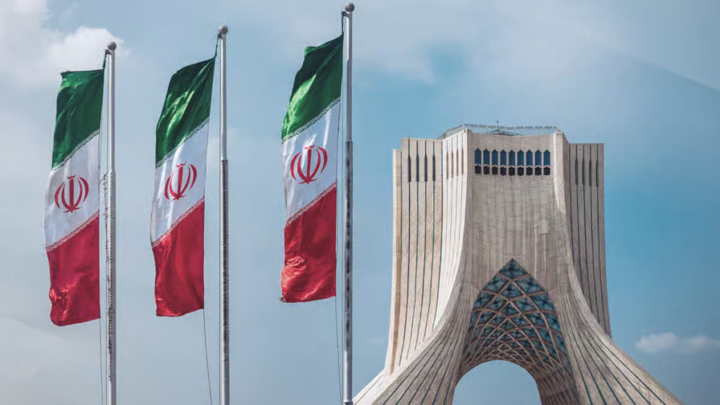Três bandeiras iranianas hasteadas ao lado da Torre Azadi em Teerã, Irã, sob um céu azul parcialmente nublado.