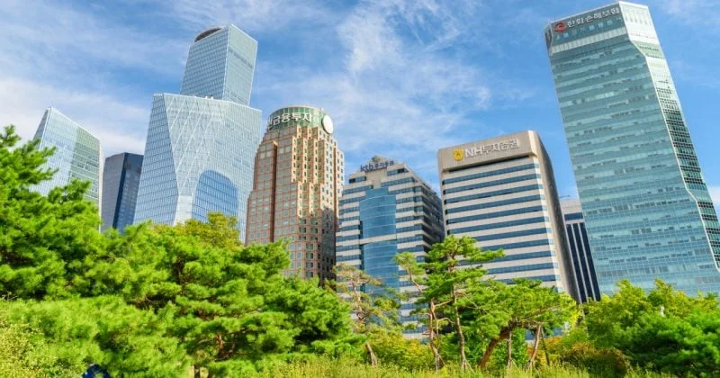 Modern skyline of Seoul's financial district featuring high-rise office towers and prominent bank headquarters, surrounded by lush green trees under a bright blue sky.