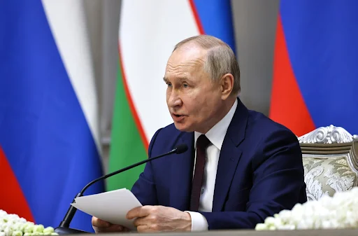 Russian President Vladimir Putin speaking into a microphone while holding papers, seated before Russian and Belarusian flags